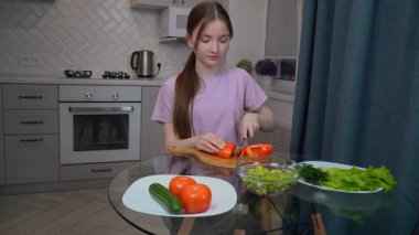 Genç kız, taze domatesler, salatalıklar, salatalıklar, marul ve dereotlarıyla çevrili, modern mutfak ortamında sağlıklı beslenme alışkanlıklarını somutlaştıran ahşap bir kesme tahtasına kırmızı biber dilimliyor.
