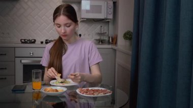 Şık bir mutfakta cam masada oturan genç bir kadın, rahat bir sabahta akıllı telefonlara bakarken hamur işlerinin ve meyve suyunun tadını çıkarıyor.