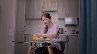 Şık bir mutfakta cam masada oturan genç bir kadın, rahat bir sabahta akıllı telefonlara bakarken hamur işlerinin ve meyve suyunun tadını çıkarıyor.