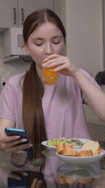 Ergen kız portakal suyunu yudumlarken besleyici salata yiyor ve yatay mutfak masasında rahat bir sabah yemeği yerken akıllı telefonlara bakıyor.