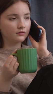 Genç kız, rahat bir şekilde mutfakta oturuyor, yeşil bardaktan çay içiyor ve telefon görüşmesi yapıyor, yüz ifadelerinin dinamik aralığını ve dikey iletişim sinyallerini gösteriyor.