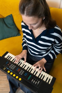 Genç bir kadın rahat sarı bir koltukta oturuyor, rahat ev ortamında müzik yapmaya kendini kaptırmış elektronik bir klavye çalıyor. Dikey fotoğraf