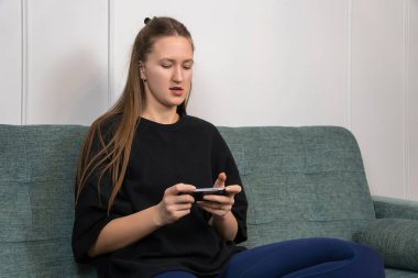 Yoğunlaşmış genç bir kadın evinde mobil oyun oynuyor rahat bir koltukta oturuyor, akıllı telefonu yatay olarak tutuyor ve iki başparmağını da kullanıyor.