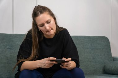 Yoğunlaşmış genç bir kadın evinde mobil oyun oynuyor rahat bir koltukta oturuyor, akıllı telefonu yatay olarak tutuyor ve iki başparmağını da kullanıyor.