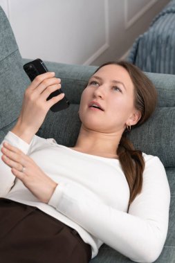 Genç bir kadın rahat bir kanepeye uzanıyor, akıllı telefon ses asistanıyla takılıyor, internette ilginç bir şey keşfederken şaşırmış bir yüz ifadesi sergiliyor. Dikey fotoğraf