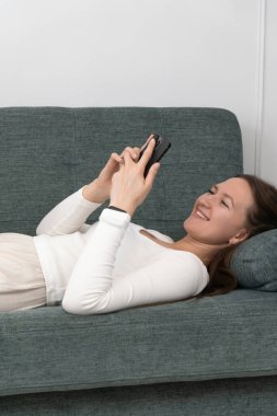 Genç kadın rahat bir şekilde koltukta uzanıyor, akıllı telefonunu huzurlu bir ifadeyle kullanıyor, modern evinde bir anlık rahatlama ve dijital bağlantının tadını çıkarıyor. Dikey fotoğraf