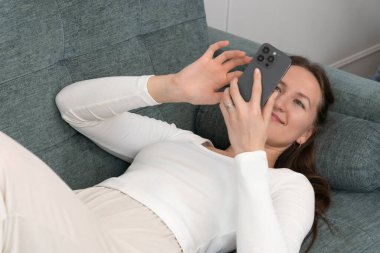 Evdeki rahat bir kanepede dinlenen genç bir kadın akıllı telefonuyla interneti ve sosyal medyayı karıştırıyor boş zamanlarının keyfini çıkartıyor.