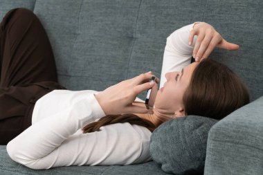 Kanepede uzanmış rahat genç bir kadın akıllı telefonunu kullanıyor, alnına eliyle dokunuyor, muhtemelen baş ağrısı ya da göz yorgunluğu geçiriyor.