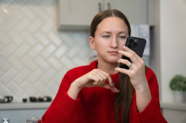 Kırmızı kazaklı genç bir kadın kahvaltıda modern bir mutfakta otururken akıllı telefonuna odaklanıyor. Rahat ve rahat bir ev ortamını işaret ediyor.
