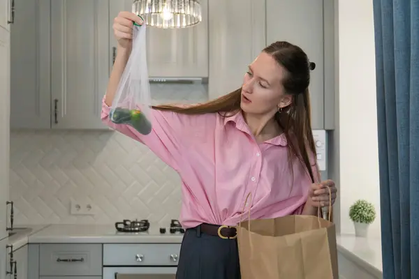 Ev hanımı elinde salatalıklı tekrar kullanılabilir bir alışveriş çantası tutuyor, modern mutfağı bozmak için teftiş ediyor, gıda atıklarının ve tüketici bilincinin altını çiziyor