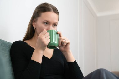 Siyah bir kazak giyen genç bir kadın rahat bir şekilde koltukta oturuyor, rahat ve davetkar bir atmosferde şık yeşil bir bardaktan gelen sıcak bir içeceğin tadını çıkarıyor.
