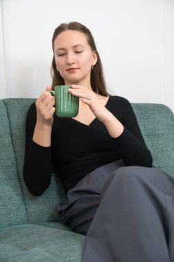 Genç bir kadın rahat bir kanepede rahatlatıcı bir anın tadını çıkarıyor, evinin rahat atmosferinde şık yeşil bir bardaktan ılık bir içeceğin tadını çıkarıyor. Dikey fotoğraf