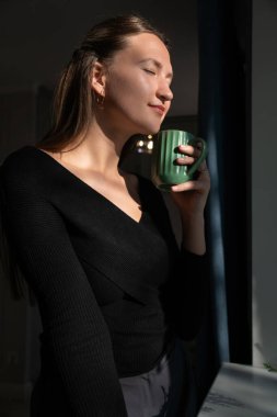 Siyah kazak giyen genç bir kadın elinde yeşil bir kupa tutuyor, küçük yeşil bir ağacı olan bir pencerenin yanında duruyor ve dairesinde güneş ışığının ve sıcak bir içeceğin tadını çıkarıyor. Dikey fotoğraf