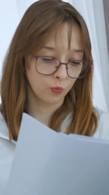 Gözlüklü ve beyaz gömlekli genç iş kadını farklı yüz ifadeleri içeren belgeler okuyor. Konsantrasyonu, şüphesi ve düşünceli dikey videoyu gösteriyor.