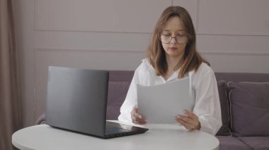 Konsantre olmuş gözlük takan ve ofis odasındaki ahşap masada çalışan, iş belgelerini dikkatle incelerken dizüstü bilgisayarda yazan beyaz gömlekli bir profesyonel.