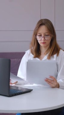 Konsantre olmuş gözlük takan ve ofis odasındaki ahşap masada çalışan, iş belgelerini dikkatle incelerken dizüstü bilgisayarda yazan beyaz gömlekli bir profesyonel.