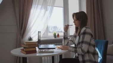 Ev ofisinden uzaktan çalışan, dizüstü bilgisayarla masada oturan, not defterine notlar yazan, doğal ışık altında kitap ve gözlüklerle çevrili, modern dijital iş yeri yaşam tarzını somutlaştıran bir kadın.