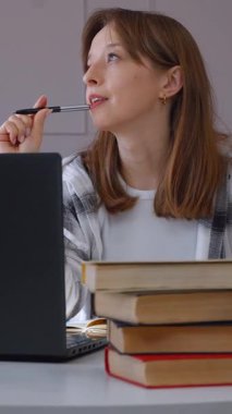 Düşünceli bir kadın ev ofis ortamında, kitap ve gözlüklerle uzaktan kumandayla çalışıyor. Odaklanmış konsantrasyon ve problem çözen dikey videolar öneriyor.