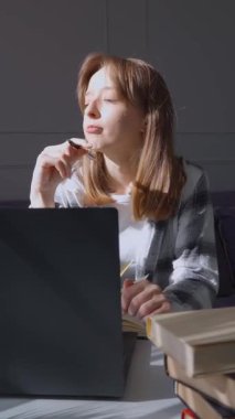 Genç bir kadın dizüstü bilgisayarını açar ve çalışmaya ya da evden çalışmaya başlar, beyaz bir masada bir yığın kitap ve dikey videosunun yanında bir bitkiyle oturur.