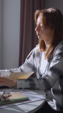 Genç bir kadın dizüstü bilgisayarını açar ve çalışmaya ya da evden çalışmaya başlar, beyaz bir masada bir yığın kitap ve dikey videosunun yanında bir bitkiyle oturur.