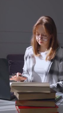 Doğal ışık, gözlük takan ve ekose gömlek giyen genç bir öğrenciyi özenle çalışırken aydınlatır. Yanında bir yığın dikey video olan dizüstü bilgisayardan notlar alır.