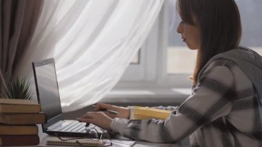 Pencereden içeri süzülen doğal güneş ışığı, bilgisayarla çalışan odaklanmış bir öğrenci ya da kadının, iş yerinde ders kitapları ve gözlüklerle çevrili el yazısıyla notlar alması.