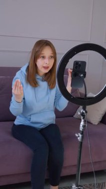 İçerik yaratan kadın video blogu kaydediyor, oturma odasındaki kanepede otururken konuşuyor ve el kol hareketi yapıyor, akıllı telefonuyla dikey video ile aydınlatılıyor.