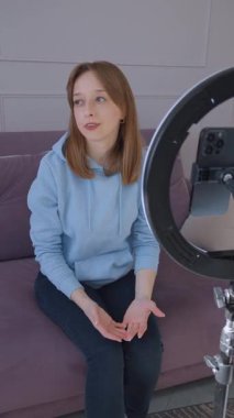 İçerik yaratan kadın video blogu kaydediyor, oturma odasındaki kanepede otururken konuşuyor ve el kol hareketi yapıyor, akıllı telefonuyla dikey video ile aydınlatılıyor.