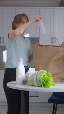 Genç kadın düzenli olarak kese kağıdından yeni alınmış paketleri açıyor, ürünleri dikkatlice mutfak masasına yerleştiriyor, organize yemek hazırlığını parlak, modern beyaz mutfak dikey videosunda sergiliyor.