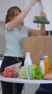 Genç bir kadın kese kağıdından paketleri boşalttıktan sonra titizlikle taze ürünleri ve bakkaliyeleri organize ediyor. Yuvarlak masa üzerinde yuvarlak, çağdaş mutfak alanında doğal ışıklandırma videosu olan şeyleri dikkatlice düzenliyor.
