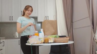 Kese kağıdından paketleri dikkatlice açan genç kadın, süt, meyve suyu, ekmek, sosis ve kurabiye gibi çeşitli yiyecek malzemelerini mutfak masasına yerleştiriyor, günlük ev yemeği hazırlama rutinini sergiliyor