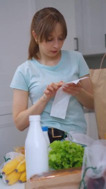 Kese kağıdından paketleri dikkatlice açan genç kadın, süt, meyve suyu, ekmek, sosis ve kurabiye gibi çeşitli yiyecek malzemelerini mutfak masasına yerleştiriyor, günlük ev yemeği hazırlama rutinini sergiliyor