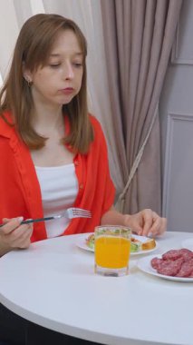 Genç bir kadın, sakin bir sabah videosu sırasında parlak modern mutfakta otururken portakal suyu, salata ve dilimlenmiş sosisle basit kahvaltıdan keyif alıyor.