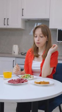 Çağdaş mutfakta masada oturan genç kadın taze salata, ekmek, salam ve portakal suyuyla besleyici kahvaltıdan keyif alıyor.