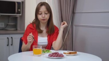 Çağdaş mutfakta masada oturan genç bir kadın taze salata, ekmek, salam ve portakal suyuyla besleyici bir kahvaltı yapıyor.