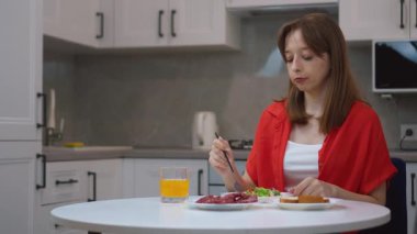 Çağdaş mutfakta masada oturan genç bir kadın taze salata, ekmek, salam ve portakal suyuyla besleyici bir kahvaltı yapıyor.