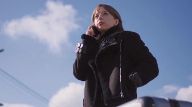 Genç iş kadını cep telefonuyla iletişim kuruyor, açık havada mavi gökyüzüne karşı duruyor dağınık bulutlarla, şık siyah ceket giyiyor, profesyonel güven yayıyor.