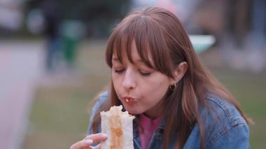 Şehir parkında bir bankta otururken baharatlı shawarma fırlatmaktan hoşlanan bir kadın öğle tatili sırasında yeşilliklerin arasında sokak yemeklerine dalmış.