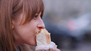 Kot ceketli bir kadın, şehir yolundaki bankta oturmuş, gevşek öğle yemeği arasında sokak yemeklerinin tadını çıkarırken Shawarma 'dan keyif alıyor.
