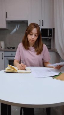 Genç bayan uzman mutfak masasında oturur, finansal belgeleri dikkatle inceler, ev bütçesini yönetirken detaylı notlar alır ve kişisel harcamaları takip eder.