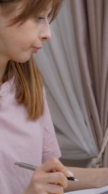 Genç profesyonel kadın aylık faturaları dikkatle gözden geçirir, kişisel çalışma alanı dikey videosundaki not defterine detaylı notlar alırken konsantre bir şekilde ev finansmanını yönetir.