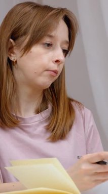 Kadın not defterine notlar yazarken dikkatli bir şekilde belgeleri gözden geçirir, iş sırasında odaklanmış konsantrasyonu gösterir veya ev ortamındaki rahat ofis ortamında dikey video çalışmasını gösterir