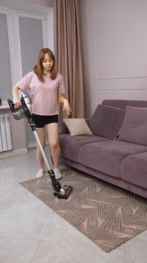 Neşeli kadın ev işi yaparken enerjik bir şekilde dans ediyor, elektrikli süpürgeyle oturma odasının halısında çağdaş kanepenin yanında dikey video kaset çekiyor.
