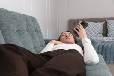 Genç bir kadın rahat bir kanepede uzanıyor, akıllı telefonunu internette gezinmek ya da video izlemek için kullanıyor, bir sesli mesaj kaydediyor, modern dairesinde huzurlu bir rahatlama anının tadını çıkarıyor.