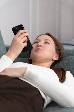 Genç bir kadın rahat bir kanepeye uzanıyor, akıllı telefon ses asistanıyla takılıyor, internette ilginç bir şey keşfederken şaşırmış bir yüz ifadesi sergiliyor. Dikey fotoğraf