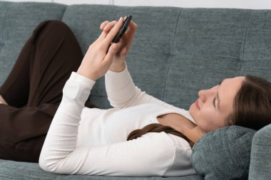 Rahat genç bir kadın kanepeye uzanıyor, akıllı telefonunu sosyal medyada gezinmek ve arkadaşlarıyla sohbet etmek için kullanıyor, huzurlu bir evde boş zamanlarının tadını çıkarıyor.