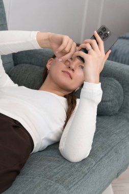 Genç bir kadın akıllı telefonunu kullanarak rahat bir kanepede uzanıyor ve parmağını ekrana doğrultuyor, sosyal medyaya bakıyor ya da internetten alışveriş yapıyor. Dikey fotoğraf