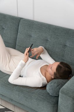 Genç bir kadın rahat bir şekilde kanepede uzanıyor, rahat giyinmiş ve akıllı telefonuna dalmış. Rahat ve rahat dairesinde huzurlu bir rahatlama ve rahatlığın tadını çıkarıyor. Dikey fotoğraf