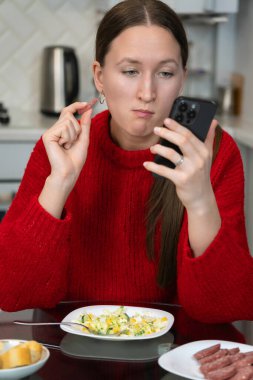 Kırmızı kazaklı genç bir kadın bir parça sosis yerken ve modern bir mutfakta akıllı telefonunu kontrol ederken gündelik ve rahat bir sabah rutinini kucaklıyor. Dikey fotoğraf