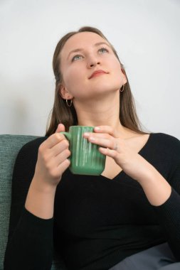 Kanepede rahat bir şekilde oturan genç bir kadın, elinde yeşil bir kupayla, düşünceli bir tefekküre dalmış, huzur ve huzur dolu bir anın tadını çıkarıyor. Dikey fotoğraf